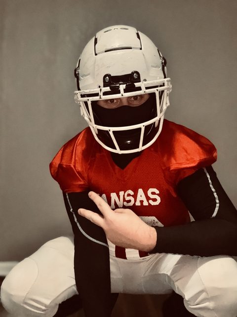 A man wearing a full American football kit, including a helmet and black balaclava, squats while posing for a photo.