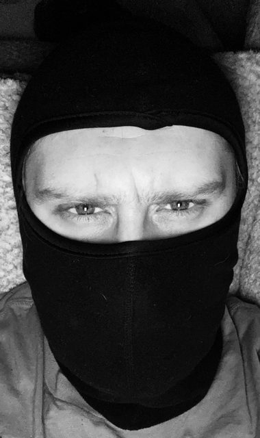 A close-up black and white portrait of a man wearing a black fabric balaclava that obscures his face while highlighting his intense gaze.
