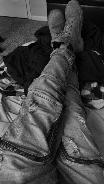 A black and white POV shot of crossed legs wearing distressed denim jeans with zipper accents and heavy work boots while lying on a bed.