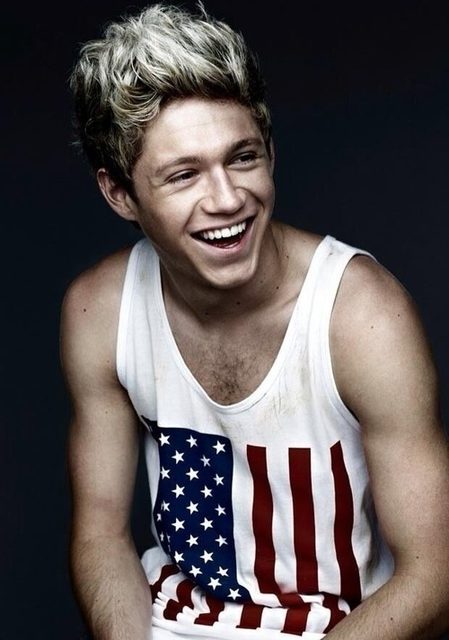 A young man with blonde highlights and a hairy chest smiles while wearing a white tank top featuring an American flag print.