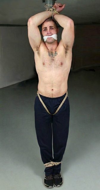 A shirtless muscular man wearing track pants is bound with ropes overhead and at the ankles, secured with a white cloth gag in a plain room.