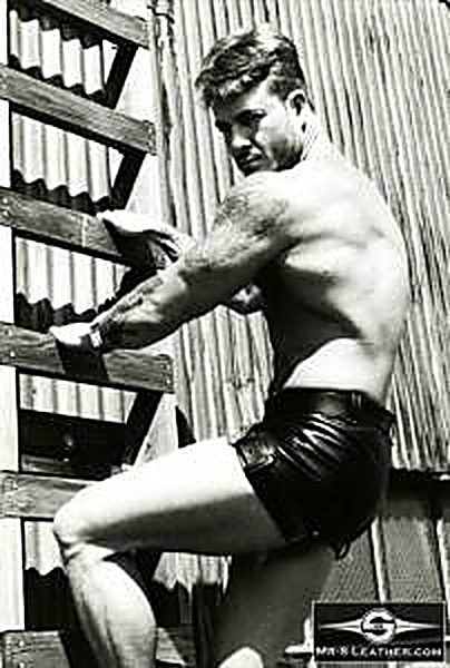 A shirtless muscular man wearing black leather shorts poses on a wooden ladder outdoors against a corrugated metal wall.