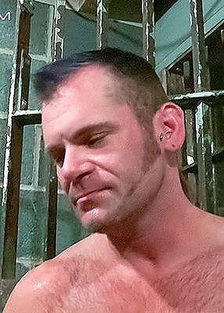 A close-up shot of a mature hairy-chested man with an ear piercing looking down in front of metal cell bars.