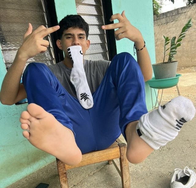 A young man sits outdoors in blue track pants, holding a tube sock in his mouth while displaying his bare foot and soles to the camera.
