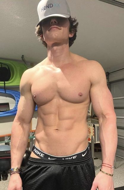 A muscular shirtless man wearing a baseball cap and gym shorts poses in a garage, showing off his defined abs and pecs.
