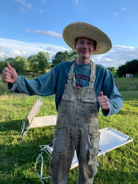 A young man in a straw hat and overalls smiles and gives a thumbs up outdoors.
