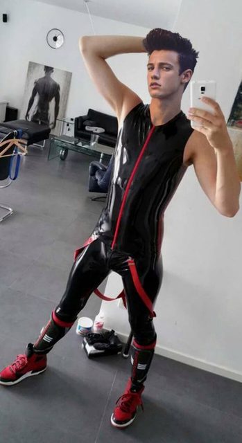 A young man in a black latex catsuit with red accents poses for a selfie indoors.