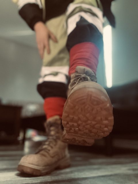A low-angle POV shot captures a person wearing tan work boots and red socks, with one boot lifted towards the viewer.