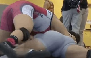 Two athletic men in tight wrestling singlets engage in a grappling match on a gym mat.