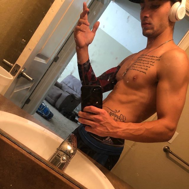A lean, muscular man with tattoos takes a shirtless mirror selfie in a bathroom wearing compression gear and headphones.