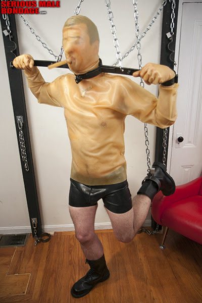 A mature man wearing a translucent latex hood and top is restrained by a spreader bar and heavy chains in a dungeon setting.
