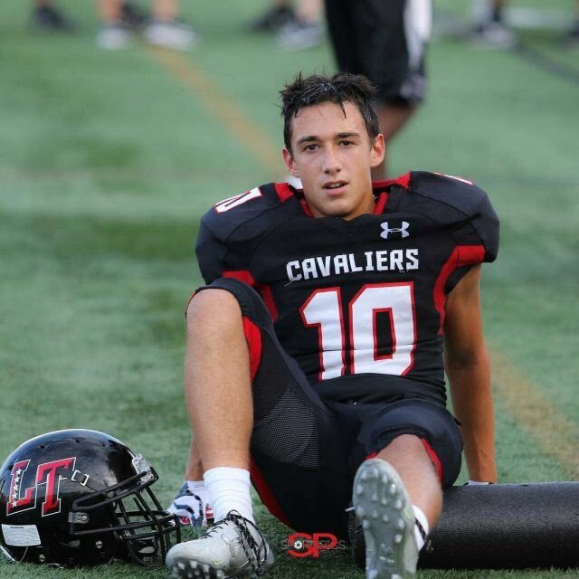 A young football player wearing a black and red Cavaliers jersey rests on the turf beside his helmet.