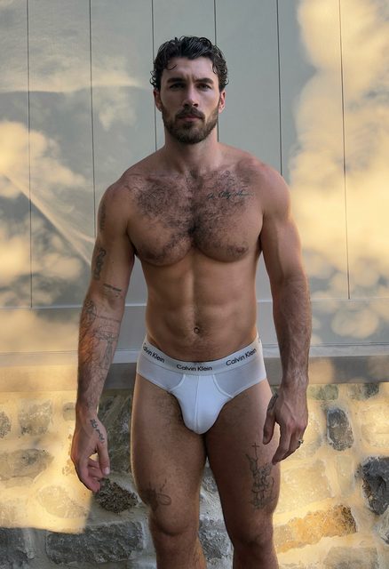 A muscular, tattooed man with a hairy chest stands outdoors wearing white briefs.