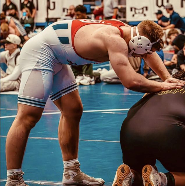A muscular wrestler wearing a tight white and red singlet and headgear leans over his opponent on a blue wrestling mat.