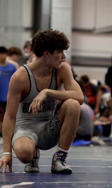 A lean, athletic young man in a grey wrestling singlet and shoes kneels on a wrestling mat during a competition.