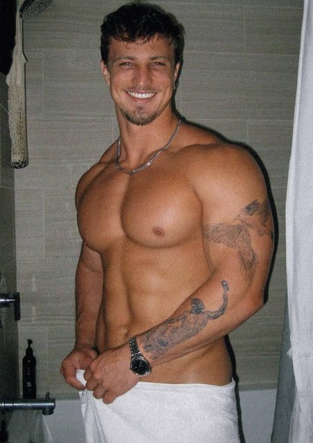 A smiling, muscular tattooed man stands shirtless in a shower holding a white towel around his waist.