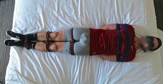 A bound and blindfolded athletic man in compression gear and socks lies tied with rope on a bed.