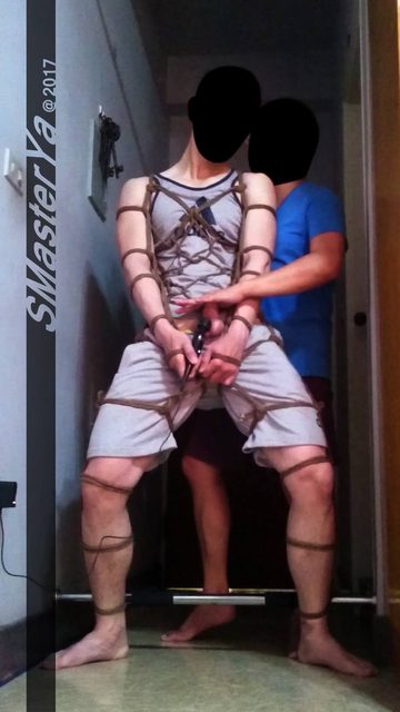 A man bound in intricate rope shibari holds a wand device while another man stands behind him.