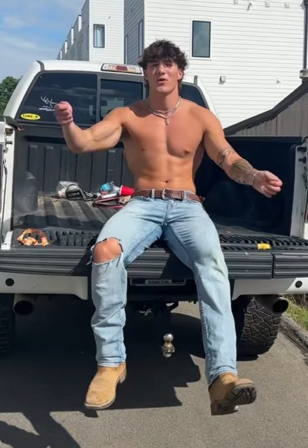 A shirtless muscular man in distressed jeans and boots sits on the tailgate of a pickup truck.