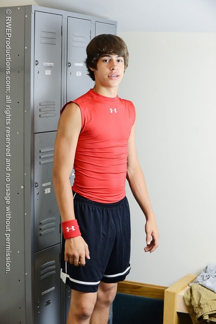 A lean athletic man wearing a red sleeveless compression top and black gym shorts stands in a locker room.