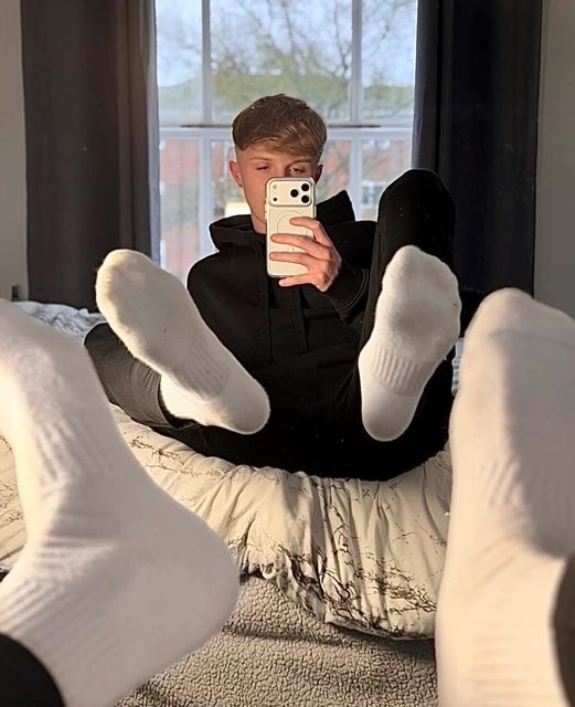 A young man in a black hoodie takes a mirror selfie while prominently displaying his feet in white socks on a bed.