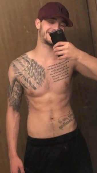 A shirtless twunk with multiple chest and arm tattoos wears a baseball cap and gym shorts while taking a mirror selfie.