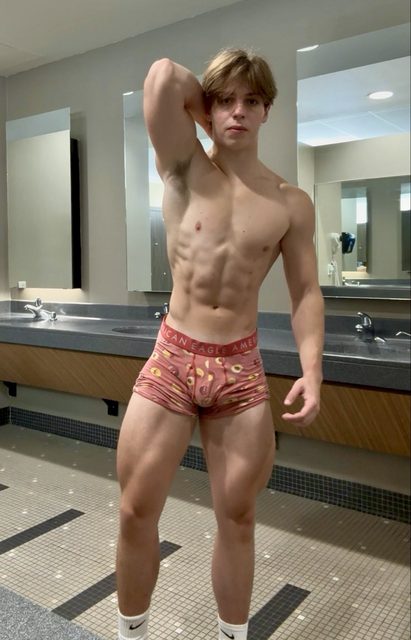 A young muscular man poses in pink patterned boxer briefs and white socks in a public locker room.