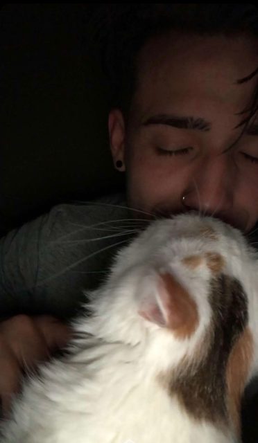 A young man with facial piercings nuzzles a calico cat in a close-up indoor shot.