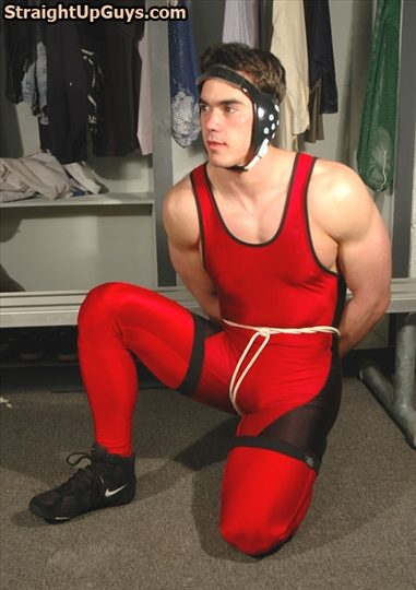 A muscular young jock in a red and black wrestling singlet and headgear kneels in a locker room with his hands tied behind his back.