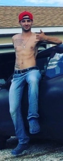 A shirtless tattooed twunk in blue jeans and a red cap leans against a car outside a garage.
