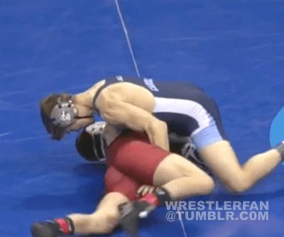 Two men in wrestling singlets and headgear are engaged in a grappling match on a blue mat.