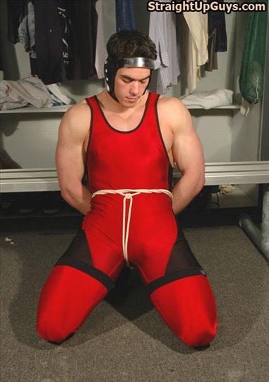A muscular man in a red wrestling singlet and headgear is kneeling in a locker room with his hands bound and ropes around his waist and groin.