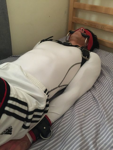 A man in a white compression shirt and gym shorts lies bound on a bed wearing a chest harness, blindfold, ring gag, and wrist cuffs.