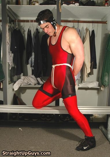 A muscular man in a red wrestling singlet and headgear stands in a locker room with his hands tied behind his back with rope.