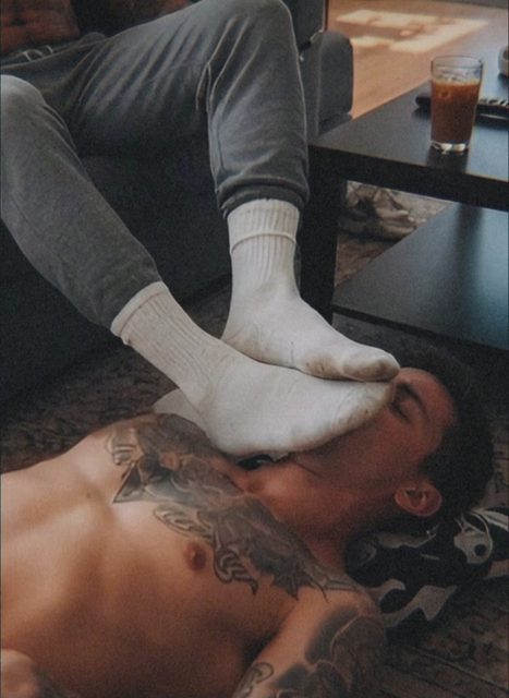 A man lying on the floor has a pair of white tube-socked feet pressed against his face by another person sitting on a sofa.