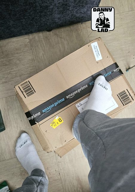 A man wearing grey sweatpants and white Dri-FIT socks rests his foot on an Amazon shipping box.