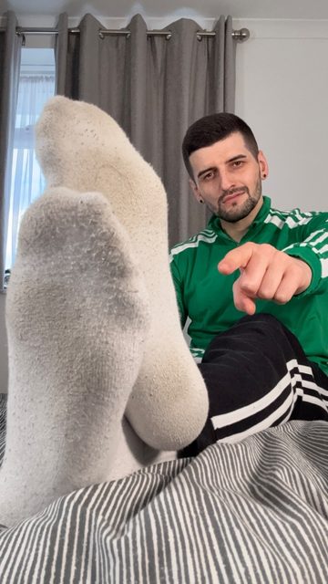 A bearded man in a green track jacket presents his white-socked feet to the camera while pointing at the viewer from a bed.