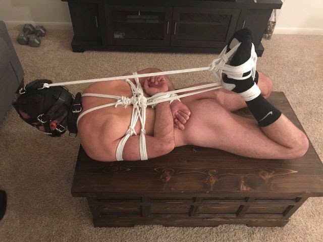 A muscular man is hogtied with white shibari rope while wearing a black leather hood and muzzle on a wooden chest.