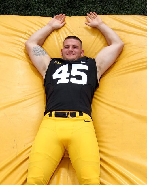 An athletic man wearing a black and yellow football uniform lies on his back on a yellow pad with his arms raised above his head.