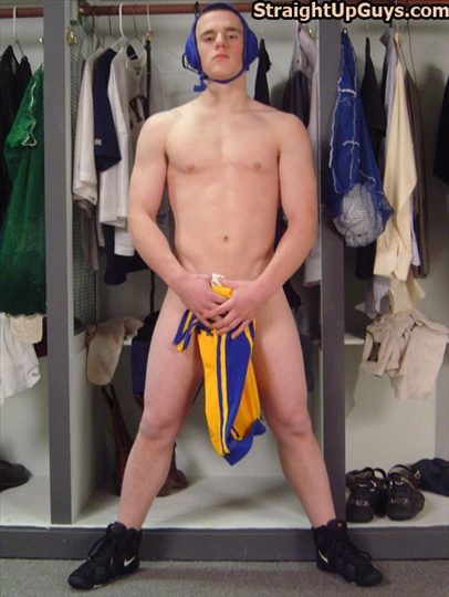 A smooth-bodied jock stands naked in a locker room wearing wrestling headgear and sneakers while holding a singlet in front of his crotch.
