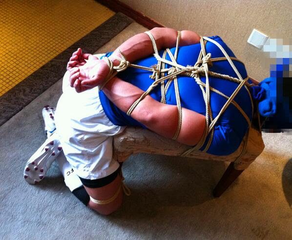 A muscular man wearing a blue sports shirt and soccer kit is securely bound in a rope harness over a bench.