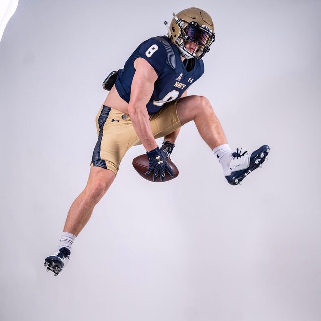 A muscular man in a Navy football uniform jumps mid-air while holding a football in a studio setting.