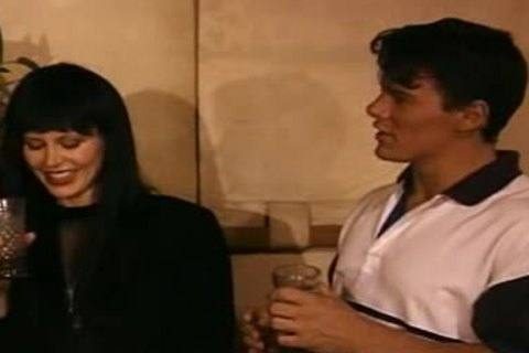 A young dark-haired man in a white polo shirt holds a glass while talking indoors with a woman.