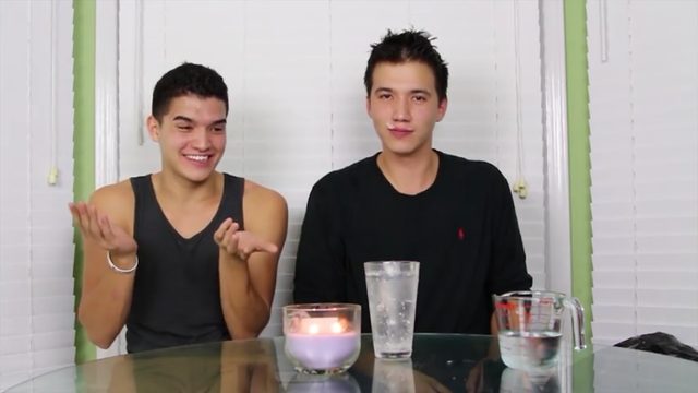 Two young, smooth-skinned men sit behind a glass table featuring a candle and water containers.