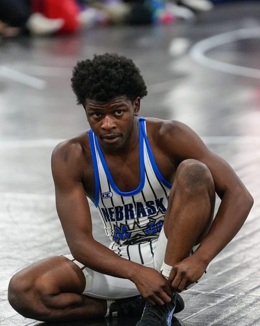 A muscular young man in a Nebraska wrestling singlet sits on a mat while adjusting his gear.