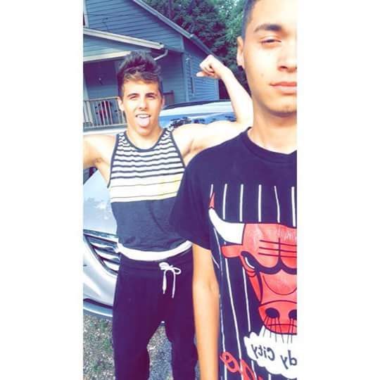 Two young men in casual sportswear pose outdoors, one flexing his bicep and sticking out his tongue.