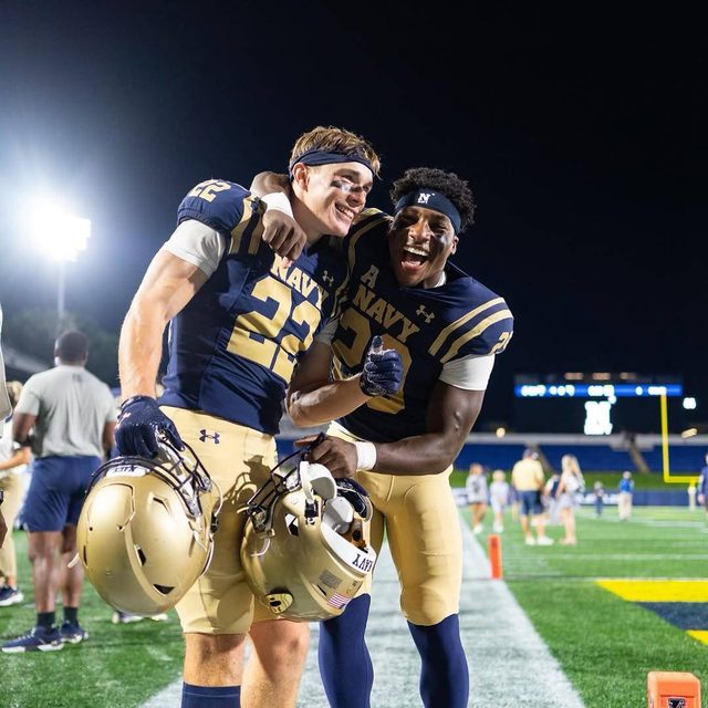 Two muscular athletes in full Navy football kits celebrate on an outdoor field at night.