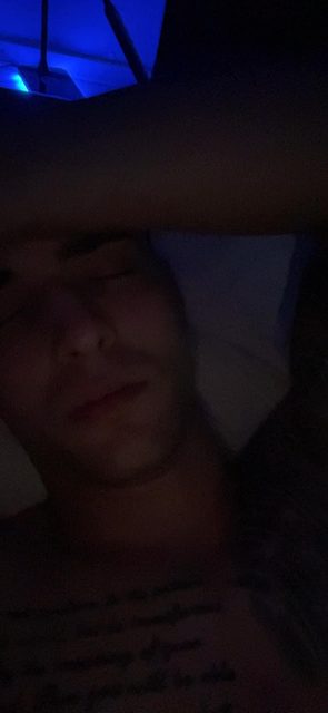 A close-up shot of a young, shirtless man with a chest tattoo lying down with his eyes closed in a dimly lit room.