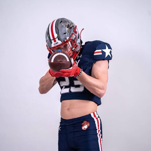 A muscular young man in a full Navy football kit with a cropped jersey poses while holding a football, revealing his toned midriff and abs.