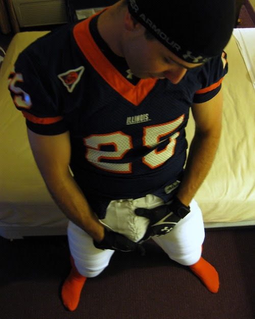 A man in a blue and orange football jersey and white padded pants poses in a bedroom while wearing gloves and holding his crotch.